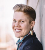 Image description: Will R. Logan, a white, nonbinary/trans person with short blond hair and blue eyes, is outside on a bright day. Will is wearing a navy sweater, plaid button-down shirt, and gold earrings. They are leaning against a wall, head turned towards the viewer, with a big, friendly smile on their face.