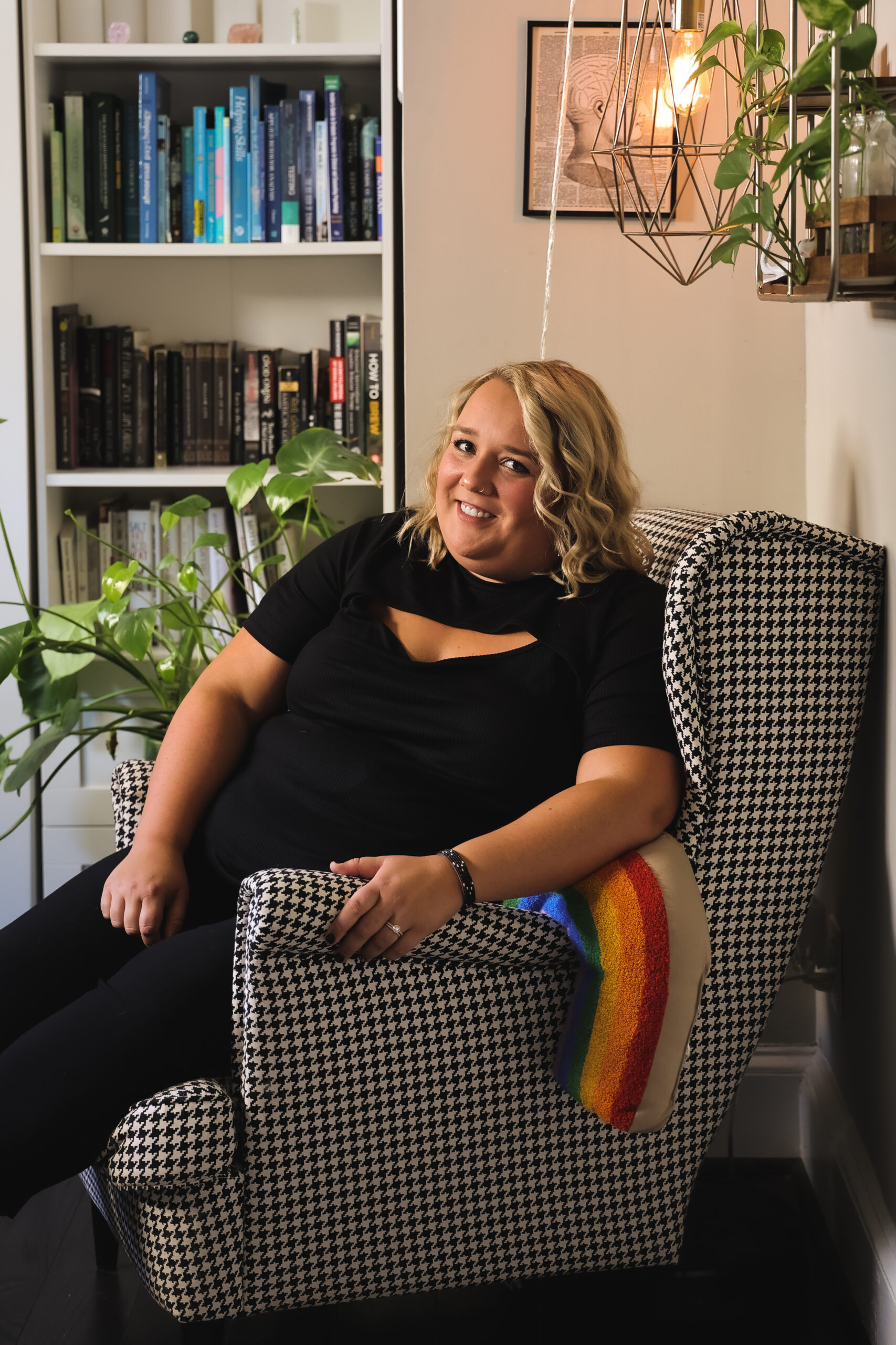 A photo of Noelle Benach, LCPC, CST - a fat, blonde woman wearing a black shirt and pants, seated in a black and white houndstooth chair with a rainbow pillow.