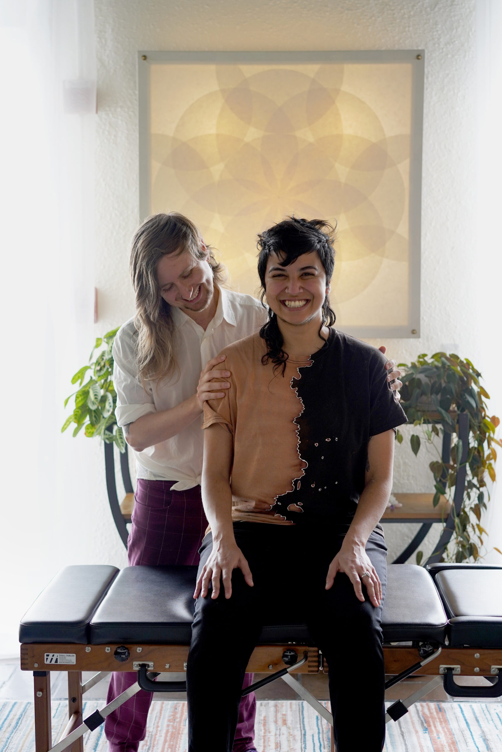 Dr. Myles, a queer chiropractor, stands behind a trans POC patient after a somatic chiropractic adjustment