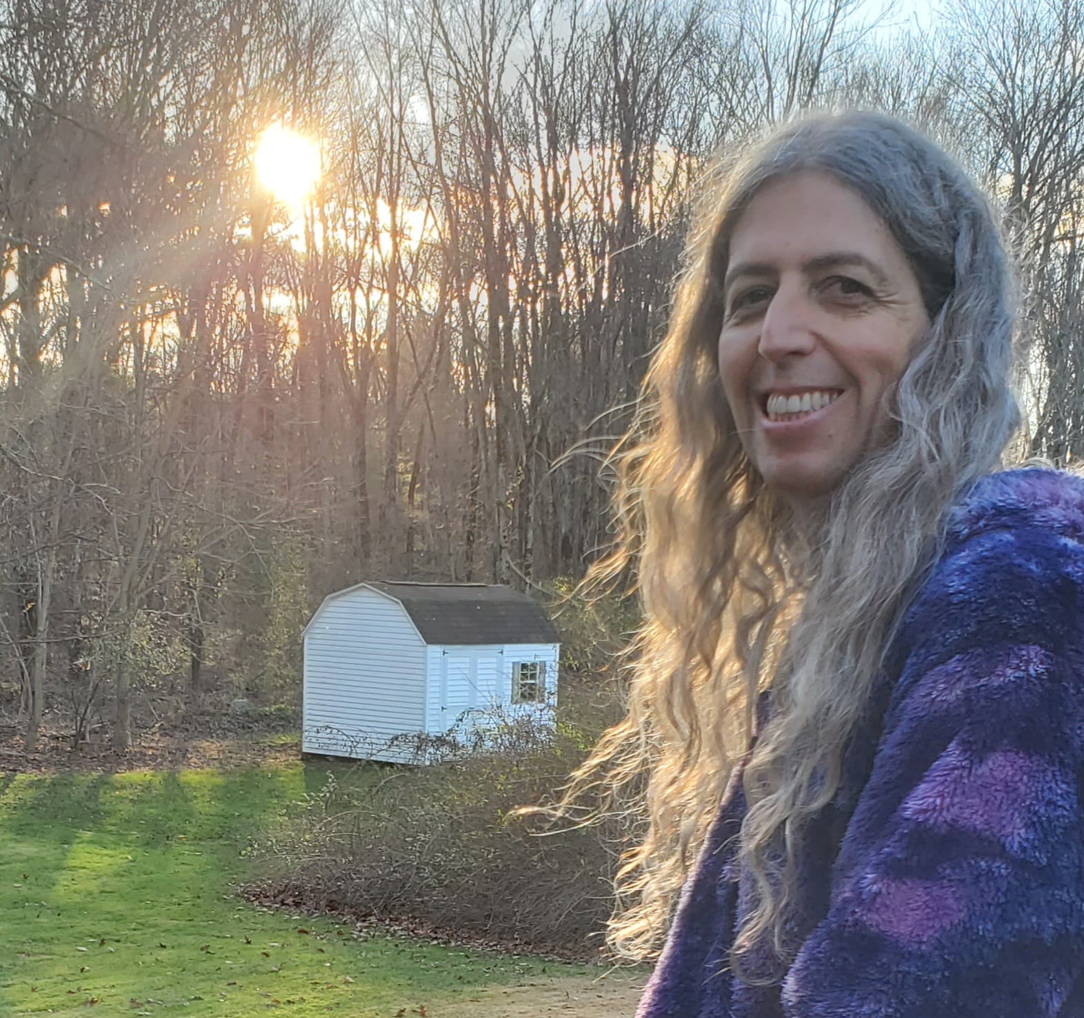 Silver haired trans woman therapist smiling against a natural background of forest & sunshine