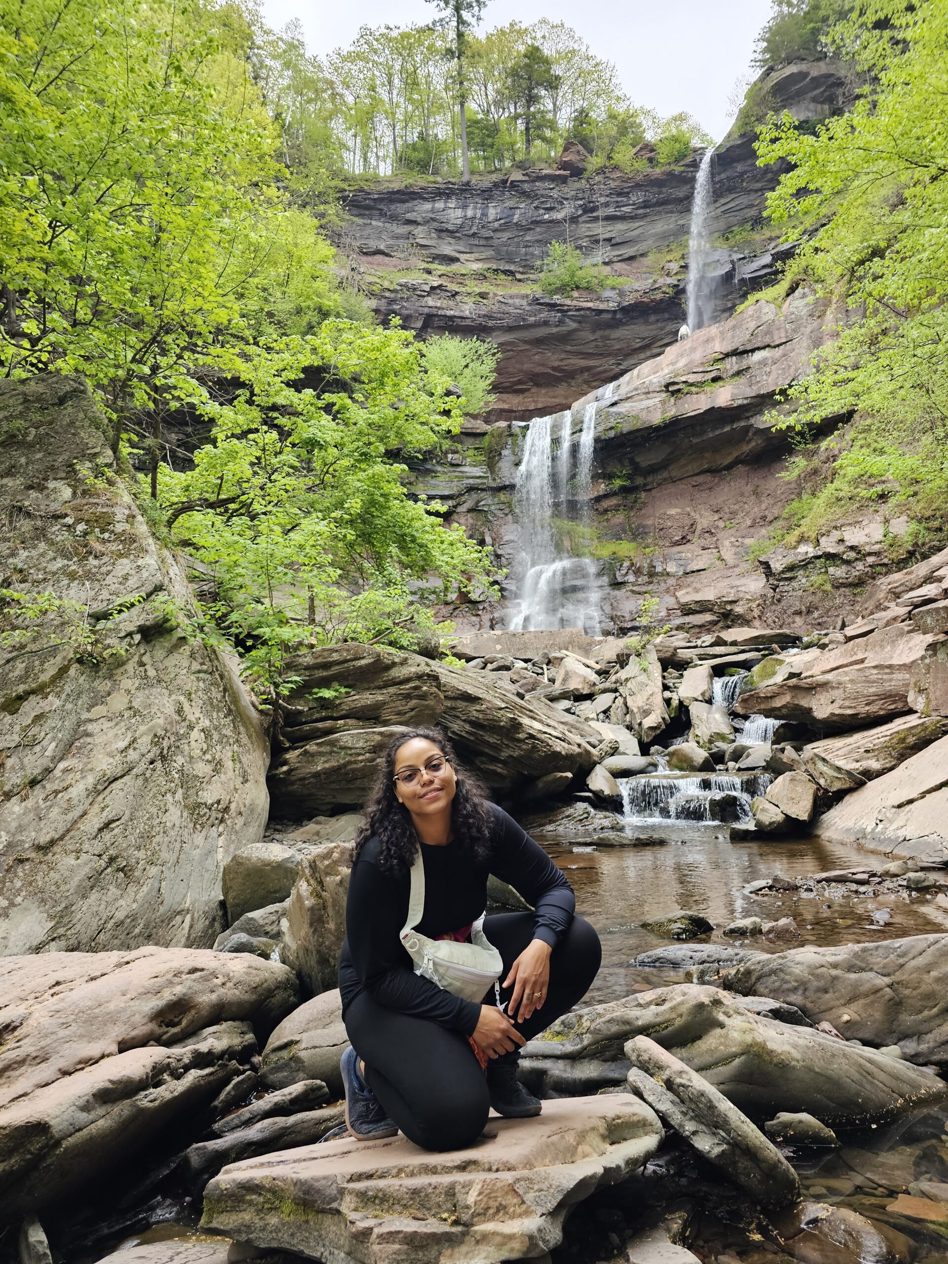 Veronica wearing glasses and black clothing, kneeling in front of a waterfall