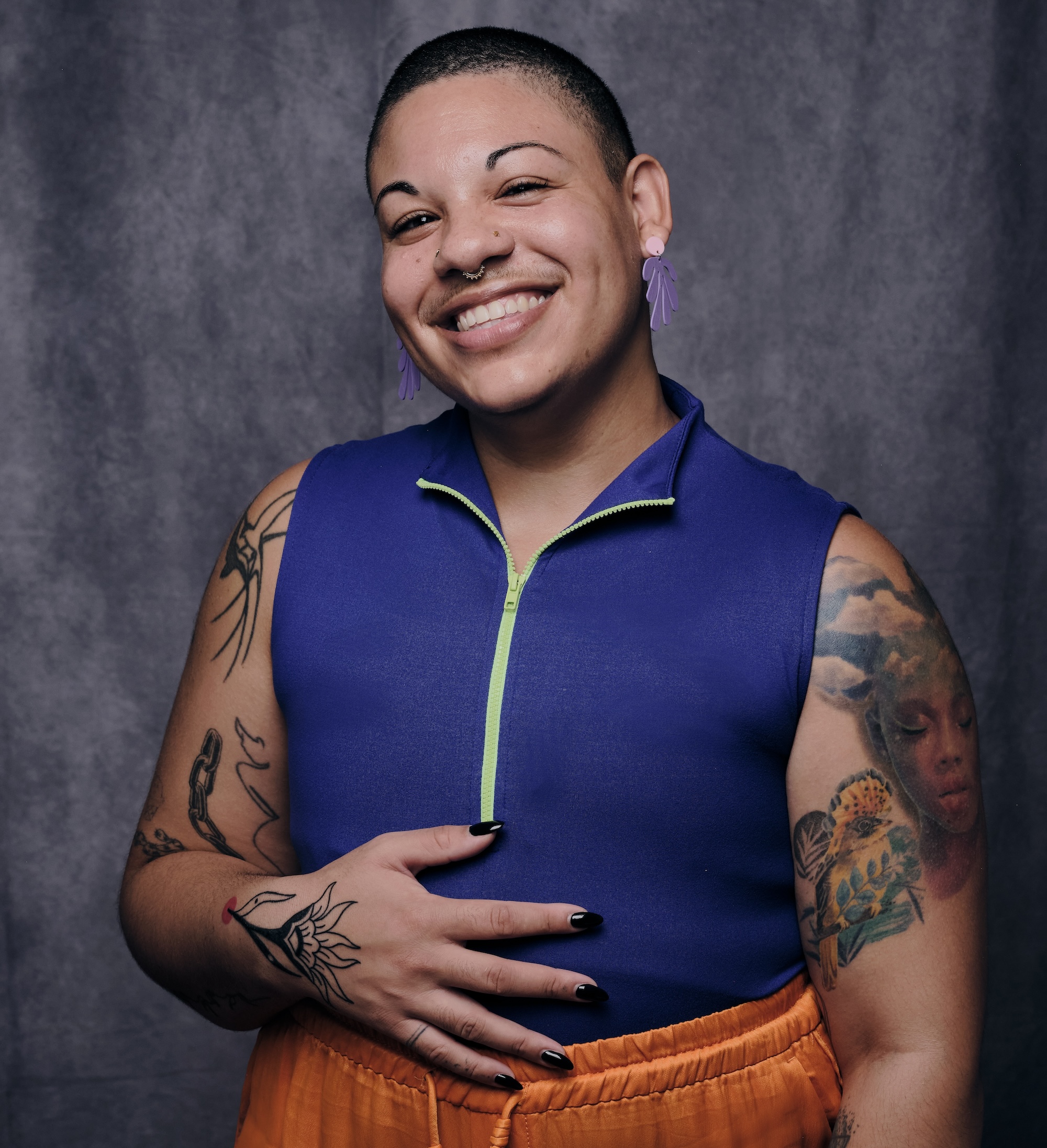 Photo of human with tan skin and a shaved head, sporting bright colored clothing, several tattoos and a smile.